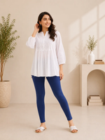 A woman modeling a white long-sleeve tunic top with delicate embroidery on the chest, paired with navy blue leggings and white slide sandals, standing in a brightly lit, minimalist beige room with potted plants and a small archway shelf.