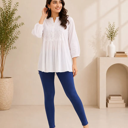 A woman modeling a white long-sleeve tunic top with delicate embroidery on the chest, paired with navy blue leggings and white slide sandals, standing in a brightly lit, minimalist beige room with potted plants and a small archway shelf.