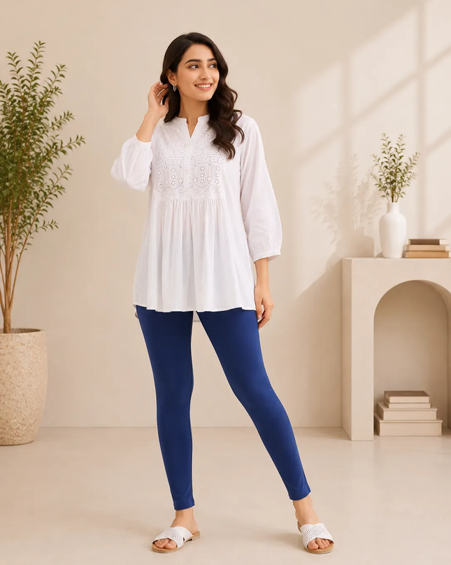 A woman modeling a white long-sleeve tunic top with delicate embroidery on the chest, paired with navy blue leggings and white slide sandals, standing in a brightly lit, minimalist beige room with potted plants and a small archway shelf.